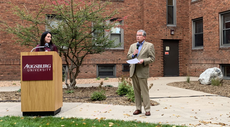 Katie Clark standing at a podium outside in the Quad as President Paul Pribbenow introduces her.